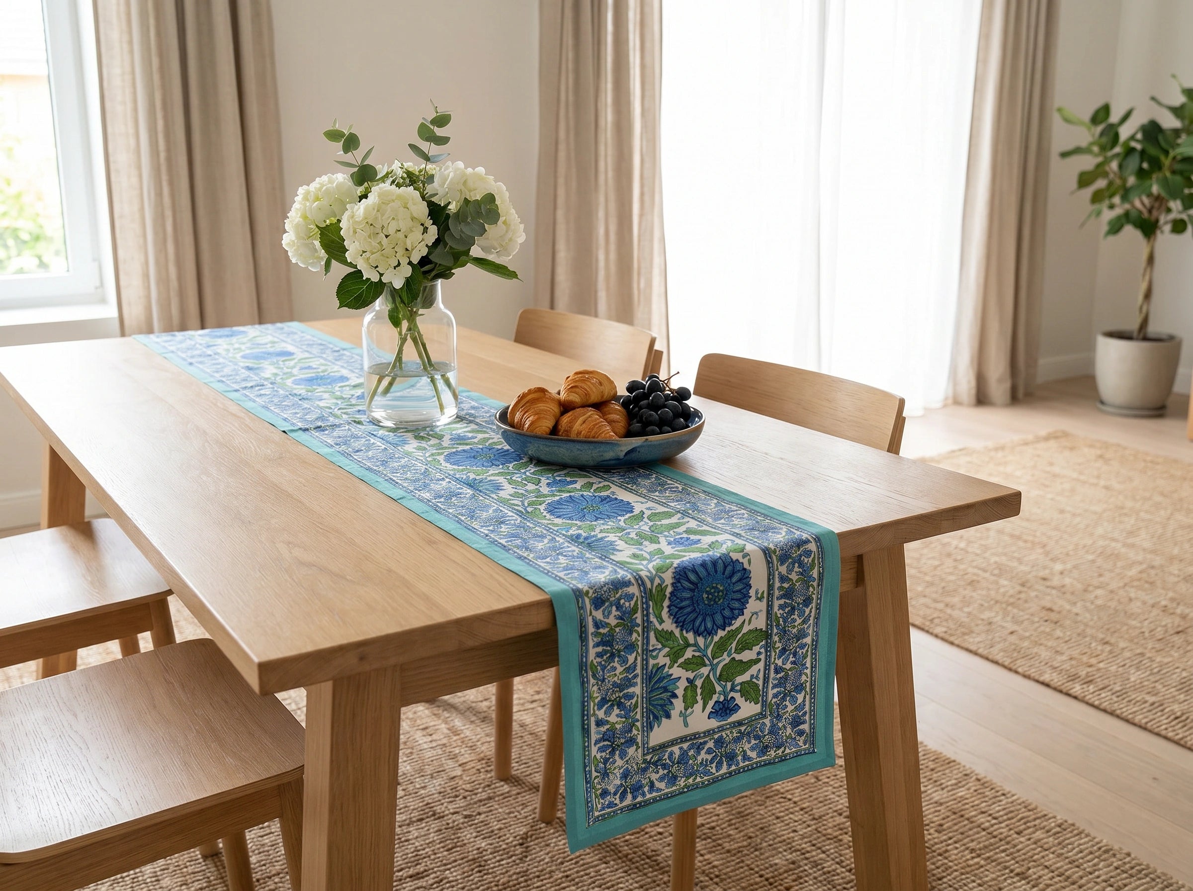 A blue and white patterned table runner with floral motifs is styled on a light wood dining table in a bright, modern room with chairs, a vase of white flowers, and a bowl of pastries.