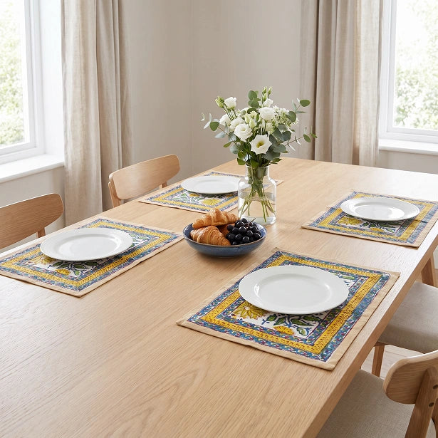 Four rectangular table placemats with a blue, yellow, and beige Mediterranean-style tile pattern are arranged on a light wood dining table. Each placemat is styled with a white plate, and the table is set with a bowl of croissants and grapes in the center and a vase of white flowers.