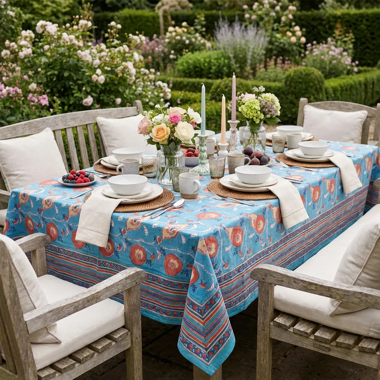 A rectangular outdoor dining table set with a turquoise blue tablecloth featuring a red and orange floral and geometric pattern. The table is surrounded by weathered wooden chairs with cream cushions, set with white plates, folded napkins, candles, and floral centerpieces, against a lush garden background.