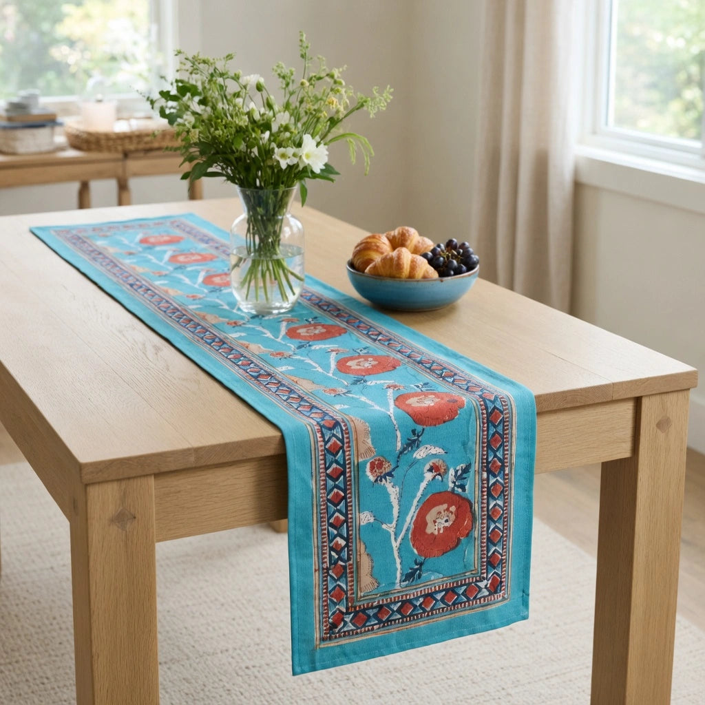 A turquoise blue table runner with red and cream floral motifs and a patterned border, styled on a light wood dining table in a bright room. A glass vase with white flowers and a blue bowl of pastries sit on the table.