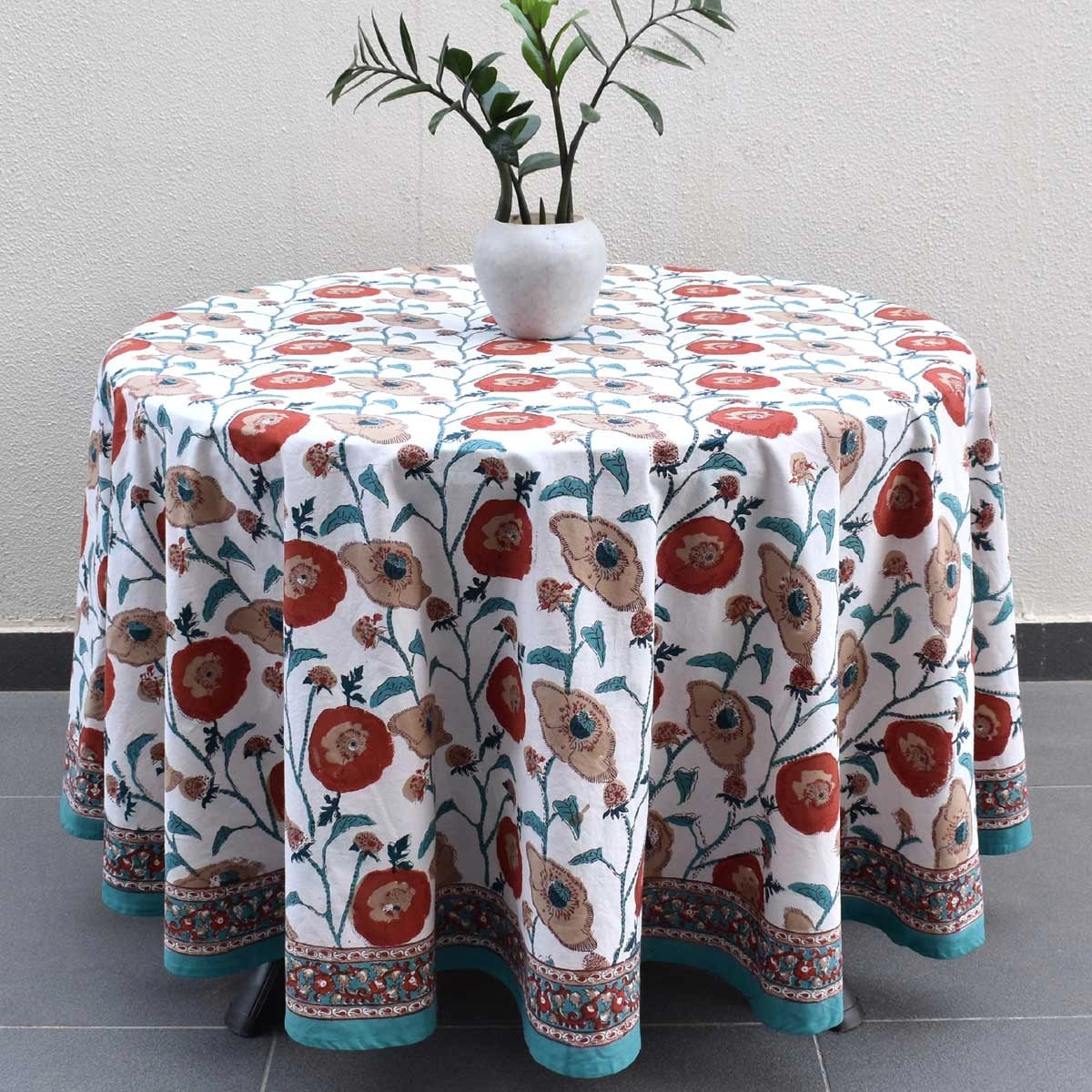 A round table covered with a white tablecloth featuring a red, teal, and beige floral pattern, styled with a white vase of green leaves on top.