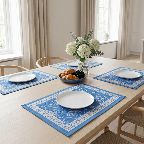 Blue and white patterned table placemats set on a light wood dining table with white plates, a floral centerpiece, and wooden chairs in a bright room.