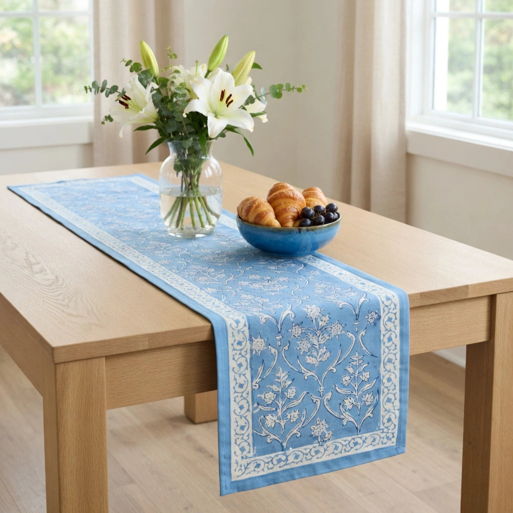 A light blue table runner with white floral embroidery and a decorative border, styled on a light wood dining table with a blue bowl of pastries and a vase of white lilies.