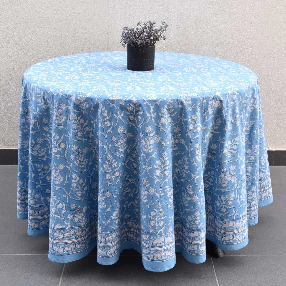 A round light blue tablecloth with a white floral and vine pattern, draped over a round table with a small black vase of dried stems on top, set against a neutral wall and tiled floor.