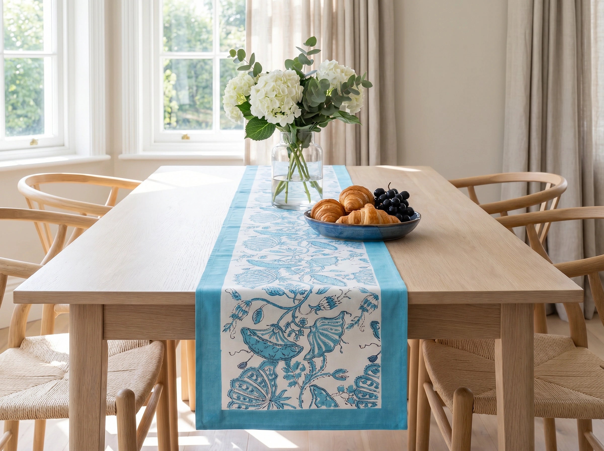 A light blue and white patterned table runner with botanical motifs is styled on a light wood dining table in a bright, modern dining room. The table runner is decorated with a blue and white patterned table runner with a botanical design, styled with a vase of white flowers and a bowl of pastries and grapes.