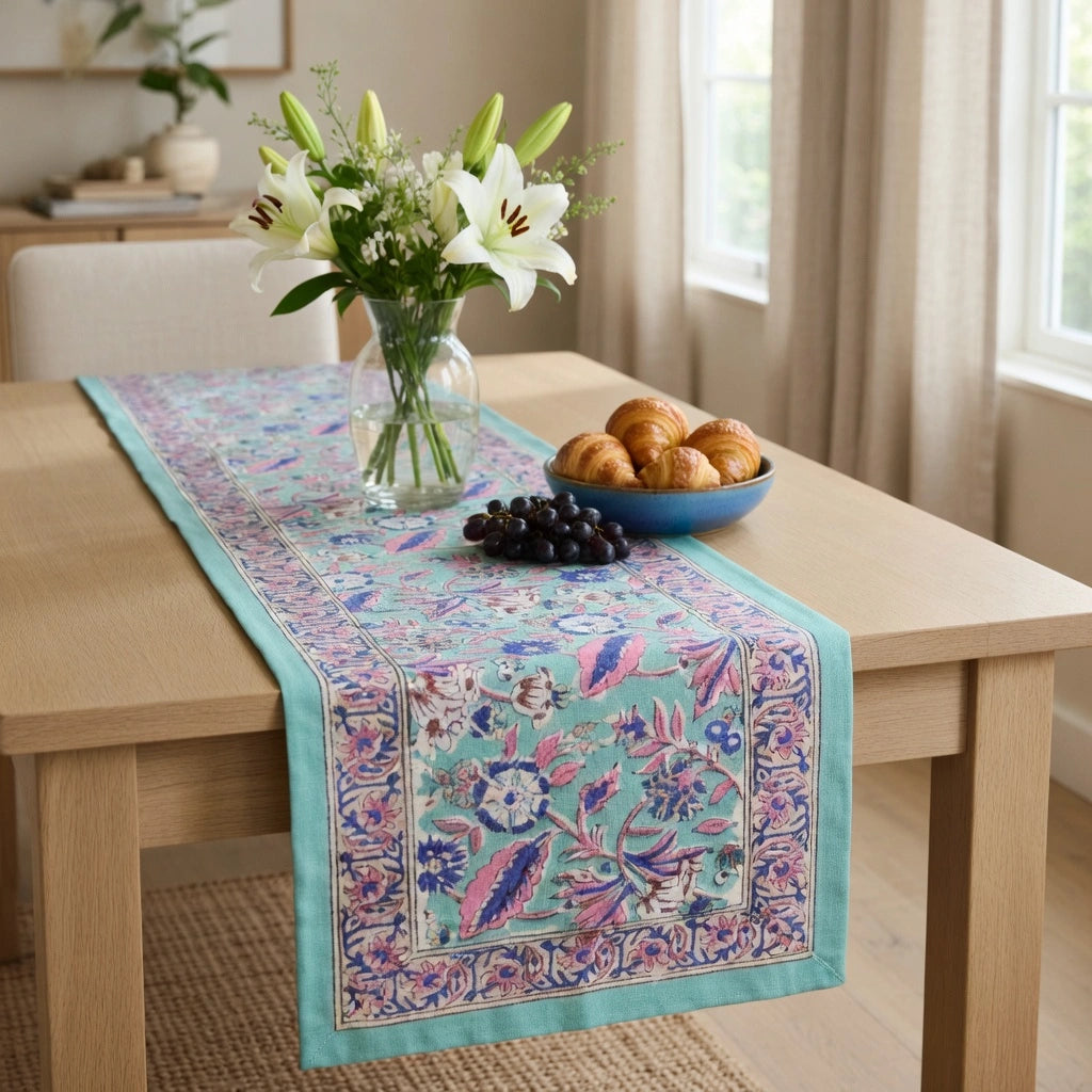 A rectangular table runner with a turquoise blue base and intricate floral and botanical patterns in pink, purple, and green, styled on a light wood dining table with a glass vase of lilies, a bowl of pastries, and a small bowl of dark grapes.