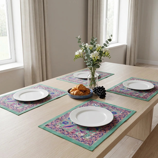Four rectangular table placemats with a colorful floral and geometric pattern in teal, pink, and purple, arranged on a light wood dining table with white plates, a vase of flowers, and a bowl of pastries in a bright room.
