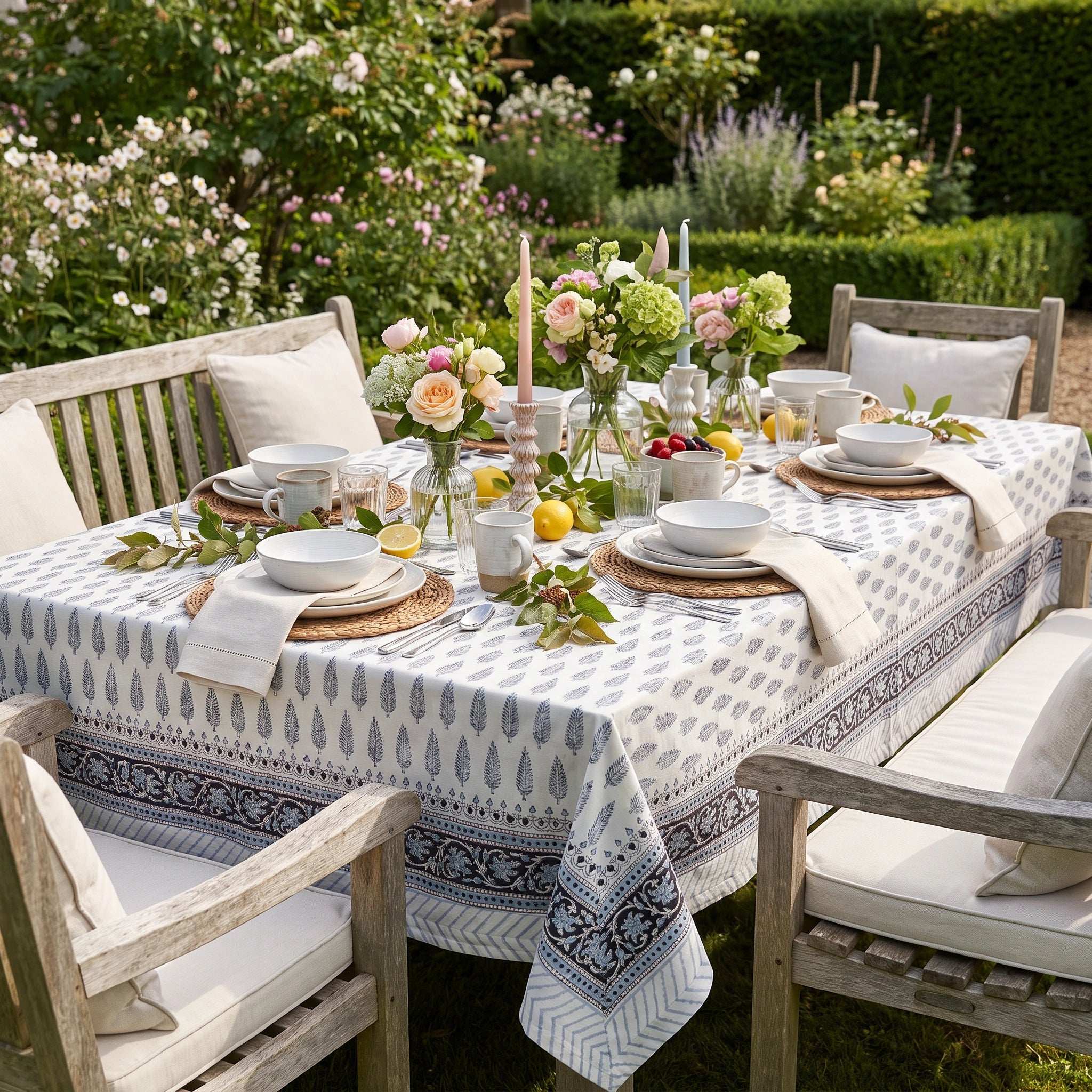 A rectangular outdoor dining table set in a garden with a white tablecloth featuring a blue geometric and floral pattern. The table is styled with white ceramic bowls, plates, folded napkins, woven placemats, floral centerpieces, and yellow lemons, surrounded by wooden garden chairs with cream cushions.
