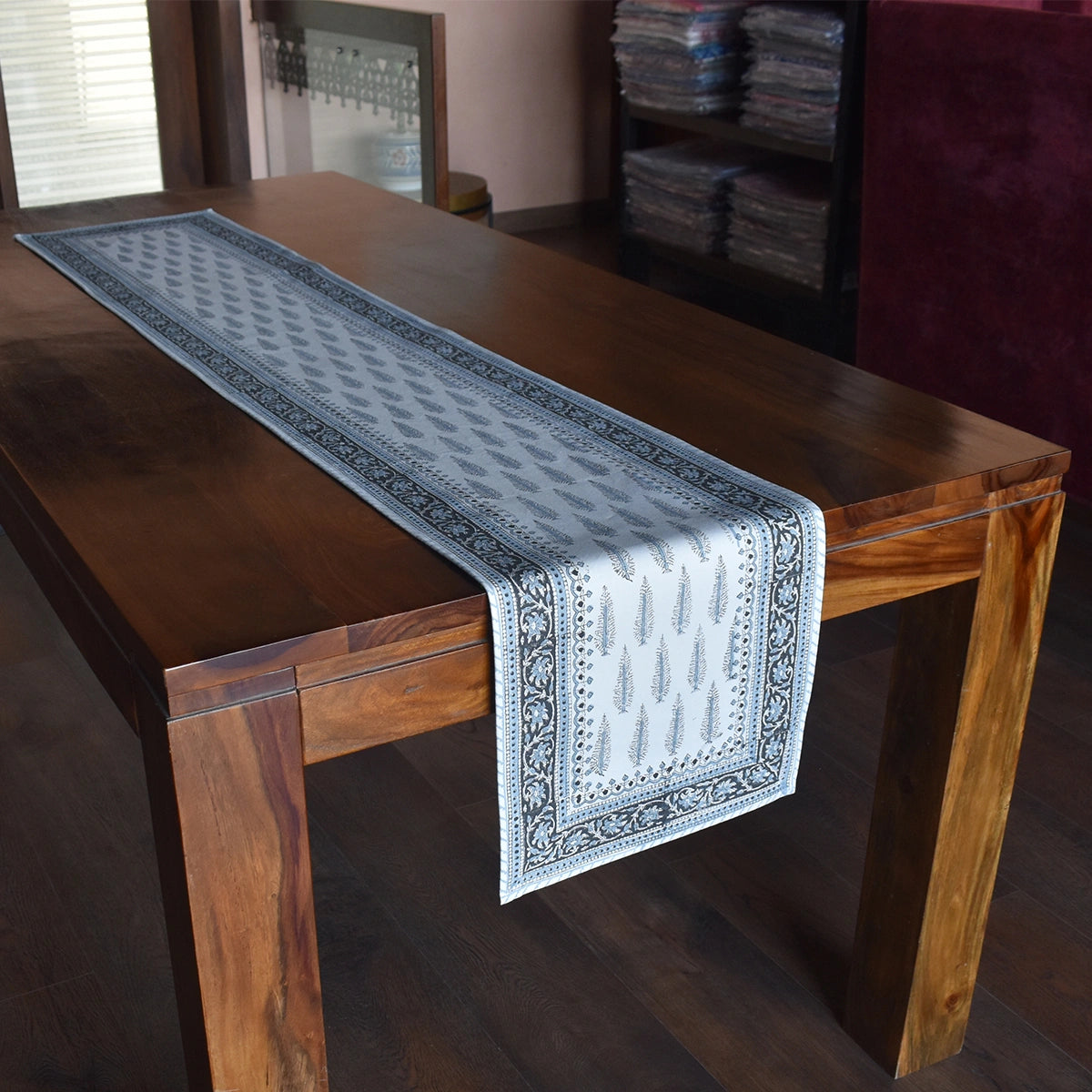 A light blue and gray patterned table runner with a black border, draped over a dark brown wooden dining table in an indoor setting.