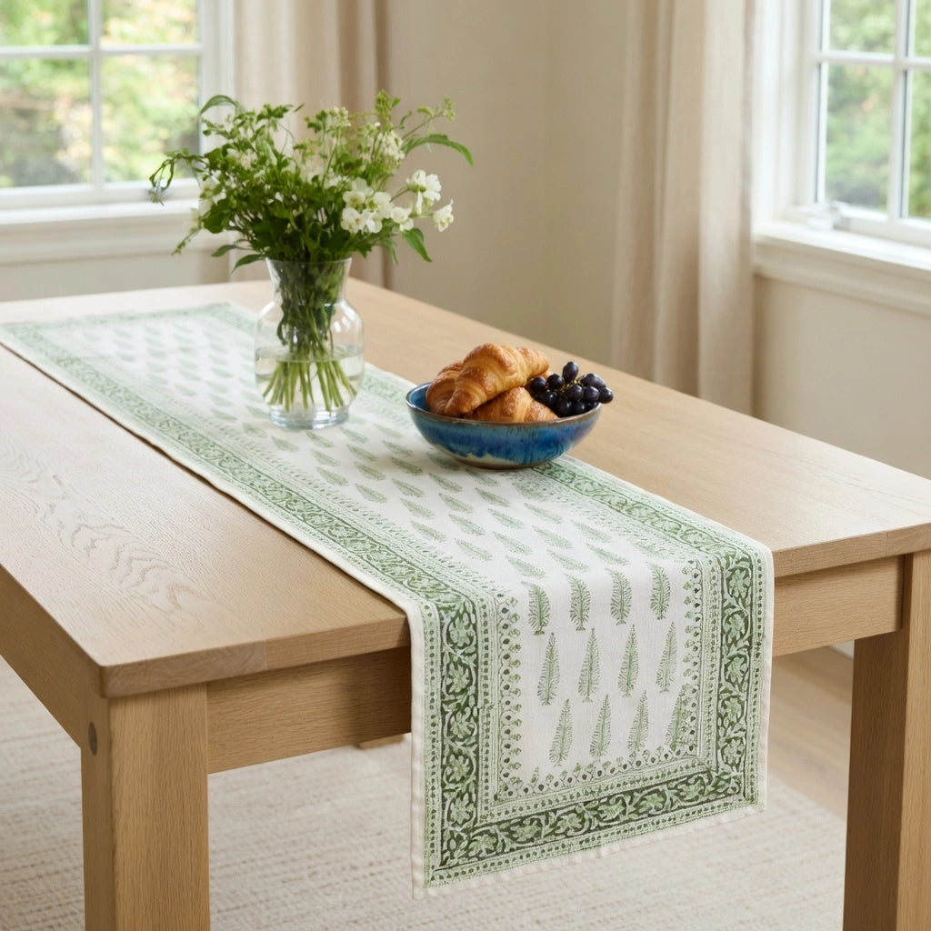 A light green and white patterned table runner draped across a light wood dining table, styled with a blue bowl of pastries and a vase of white flowers in a bright room.