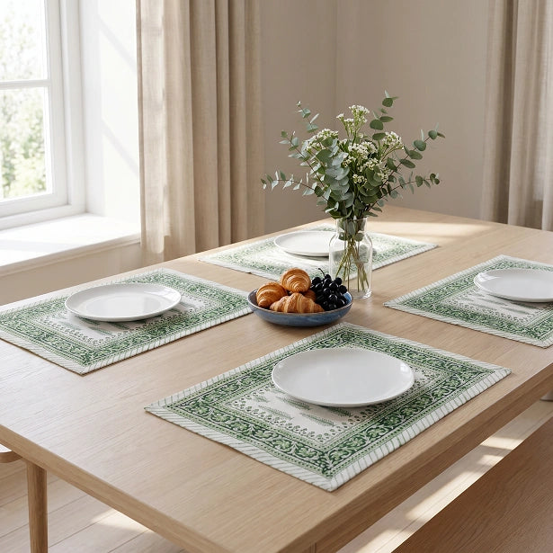 Four rectangular table placemats with a green and white traditional floral border pattern are arranged on a light wood dining table. Each placemat holds a white plate, and the table is styled with a central glass vase of greenery and a bowl of pastries and grapes in a bright, sunlit room.